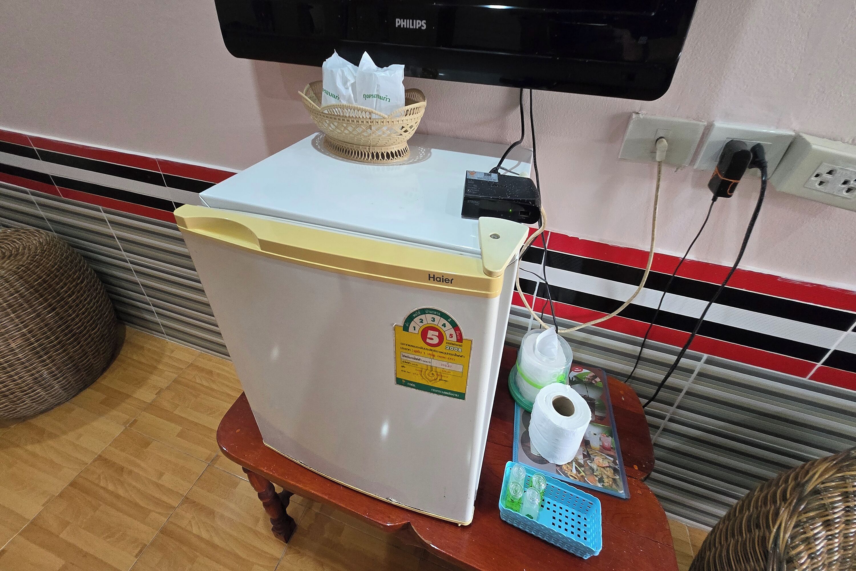 Un refrigerador blanco con una etiqueta amarilla, una cesta con pañuelos y una regleta de enchufes sobre una mesa de madera.