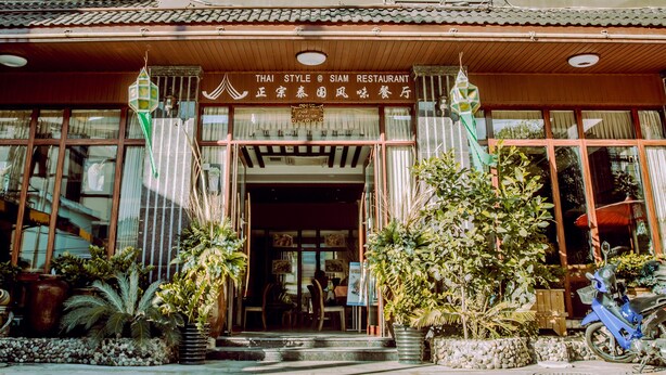 En thairestaurang med en traditionell träfasad, stora fönster och krukväxter i entrén.