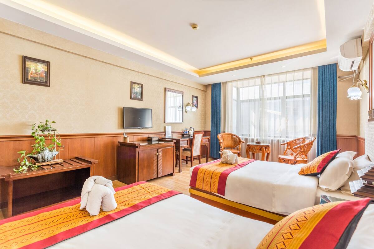 A hotel room with two beds, a desk, a TV, and a large window with curtains.