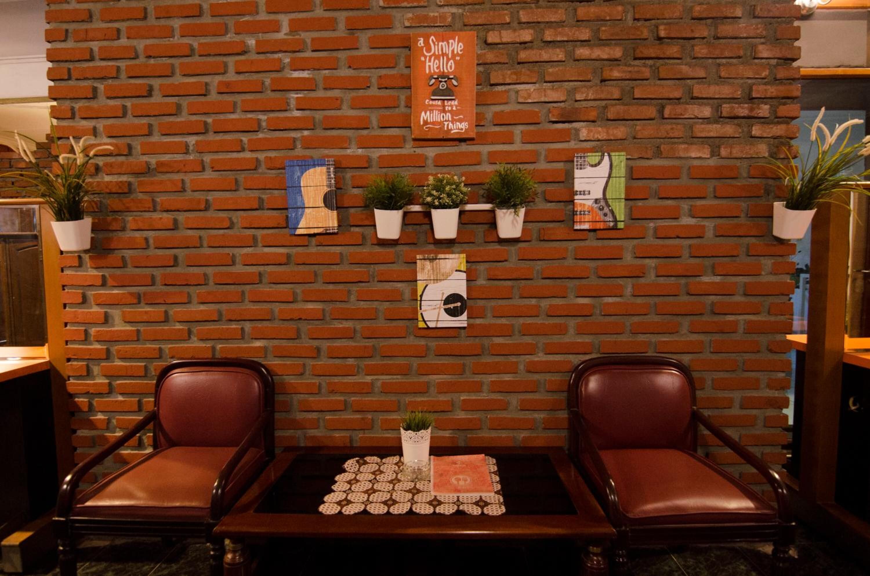 Un mur de briques orné d’une pancarte sur laquelle on peut lire « Bonjour », avec des plantes en pot et une petite table sur laquelle sont posés un set de table et un vase.
