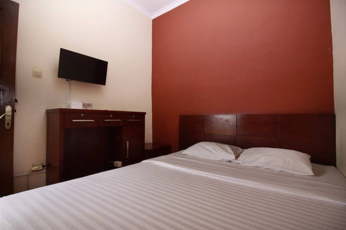 A hotel room with a bed, a TV mounted on the wall, a wooden dresser, and a red accent wall.