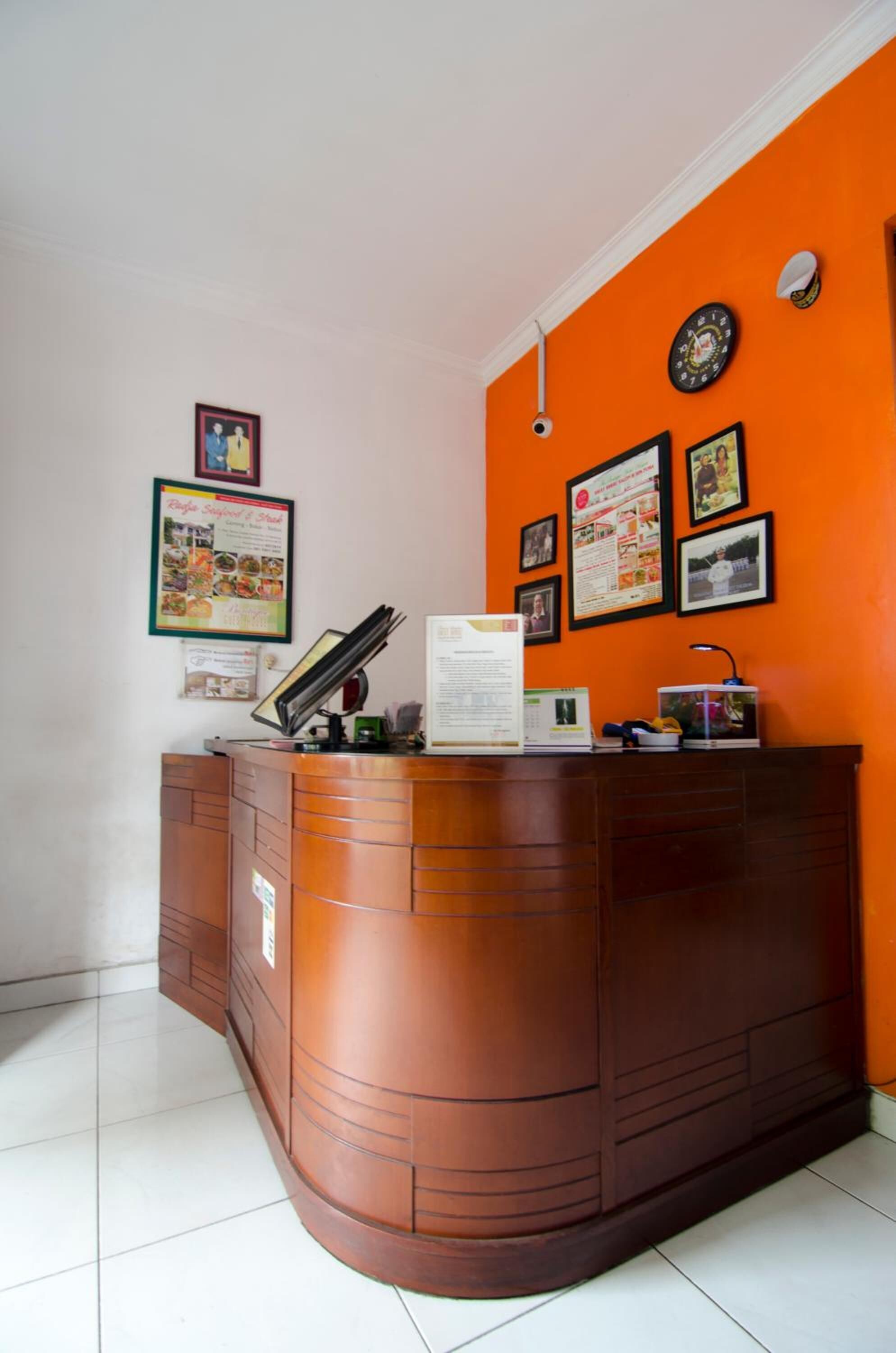 A reception desk with a wooden finish, a white tiled floor, and an orange wall with framed pictures and a clock.