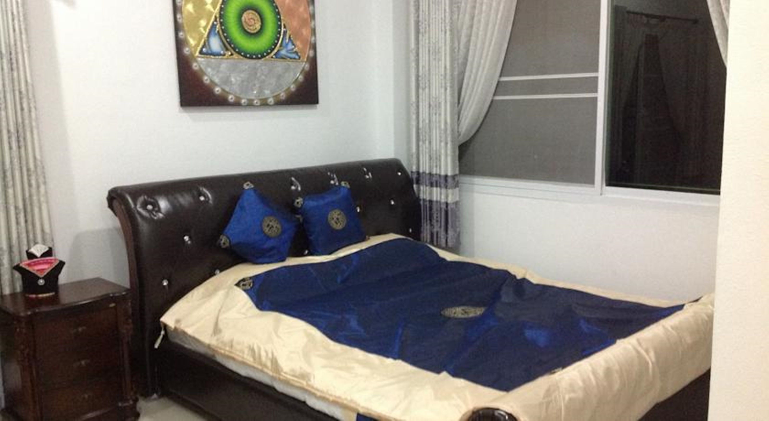 Un dormitorio con cama, mesita de noche, un adorno en la pared y una ventana con cortinas.
