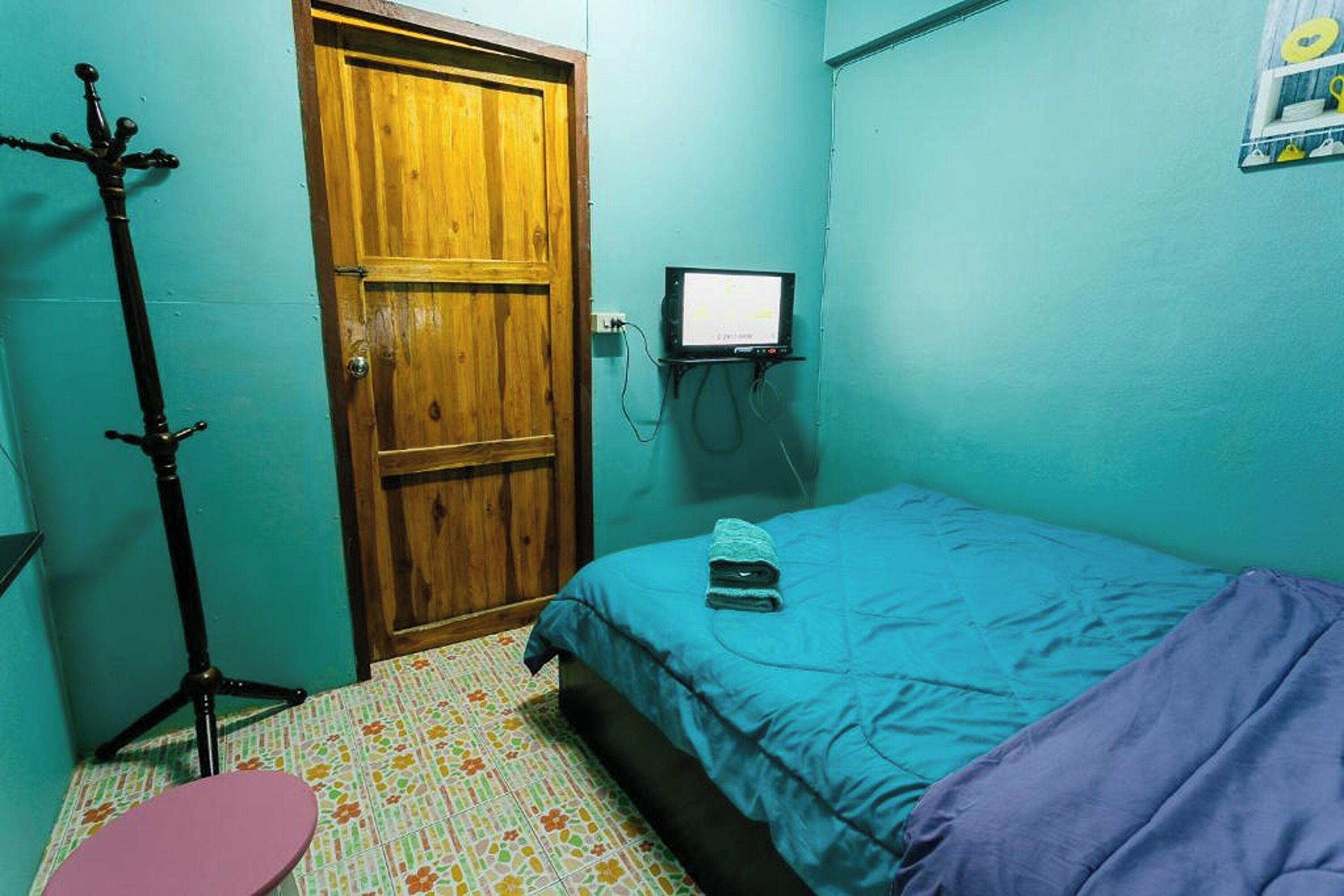A room with a wooden door, a bed with a blue blanket, a small table, and a floor lamp.