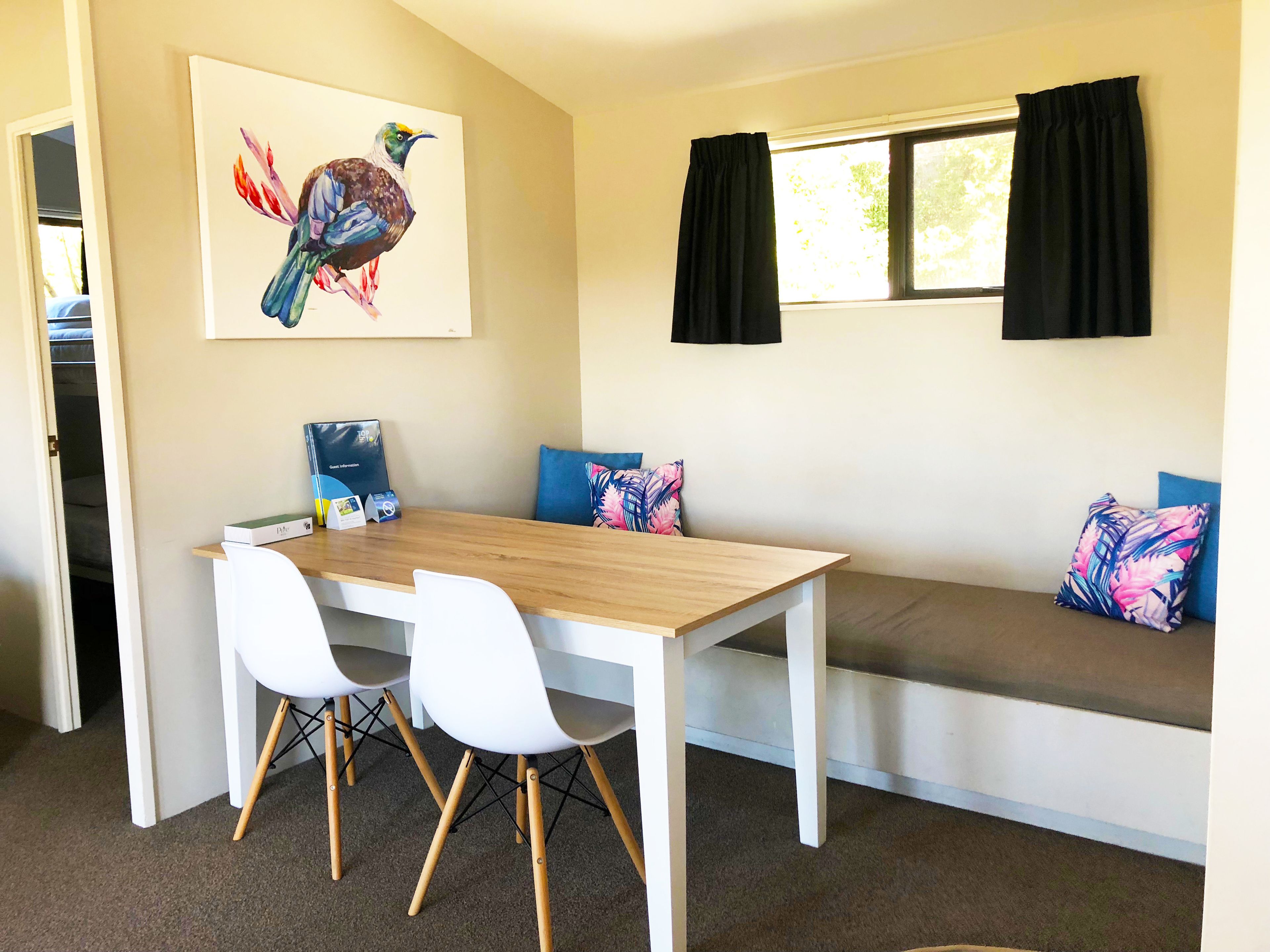 A dining area with a wooden table, white chairs, a bench with colorful cushions, a painting of a bird, and a window with black curtains.