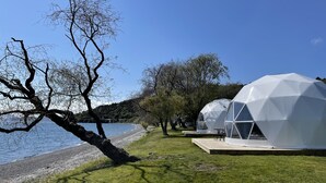A lakeside view with transparent dome-shaped accommodations, a wooden deck, and a tree-lined path. - Motutere Bay Top 10 Holiday Park (Motutere)