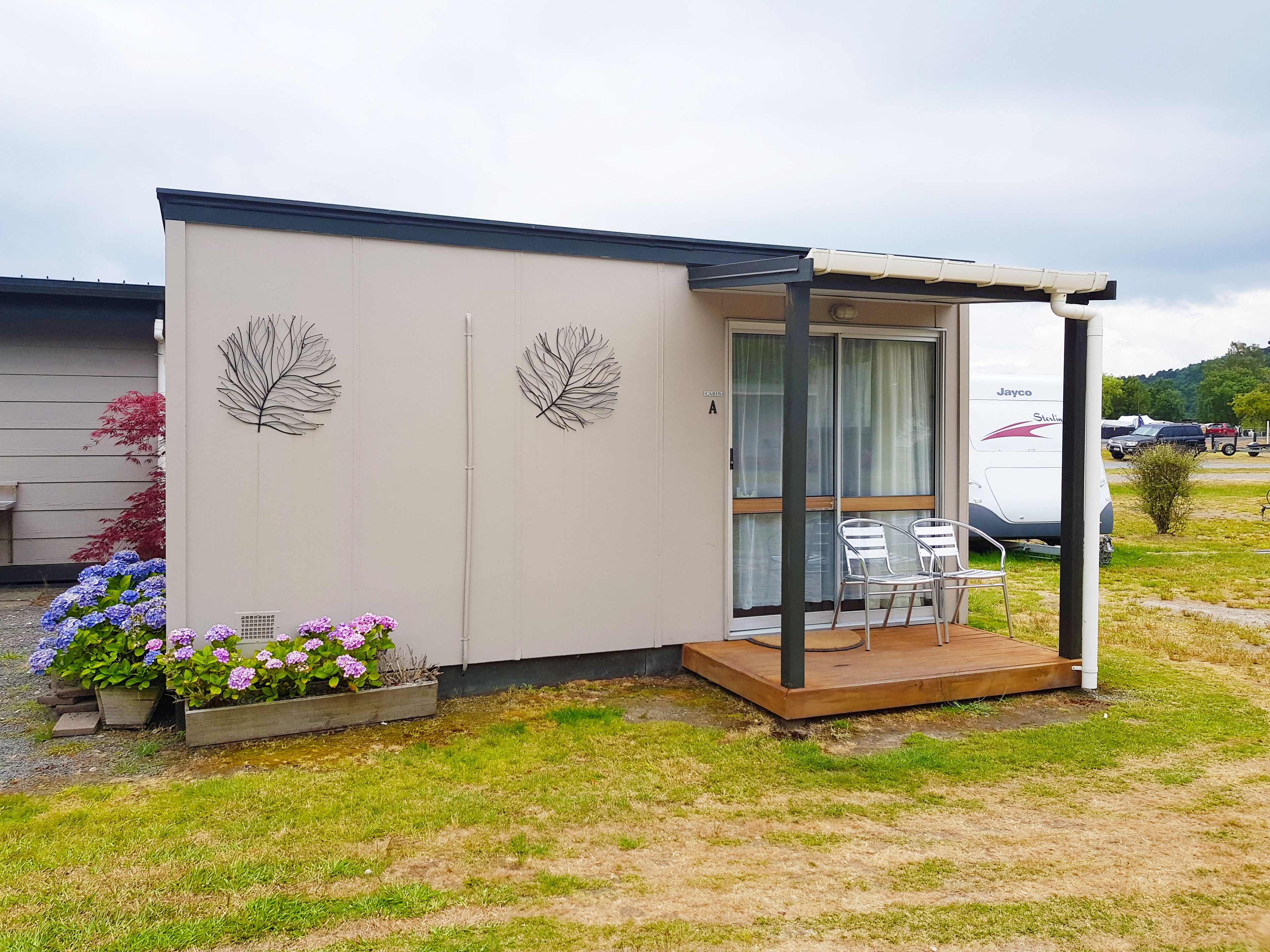 A small, modern outdoor seating area with a glass door and a wooden deck, surrounded by a garden with purple and pink flowers.