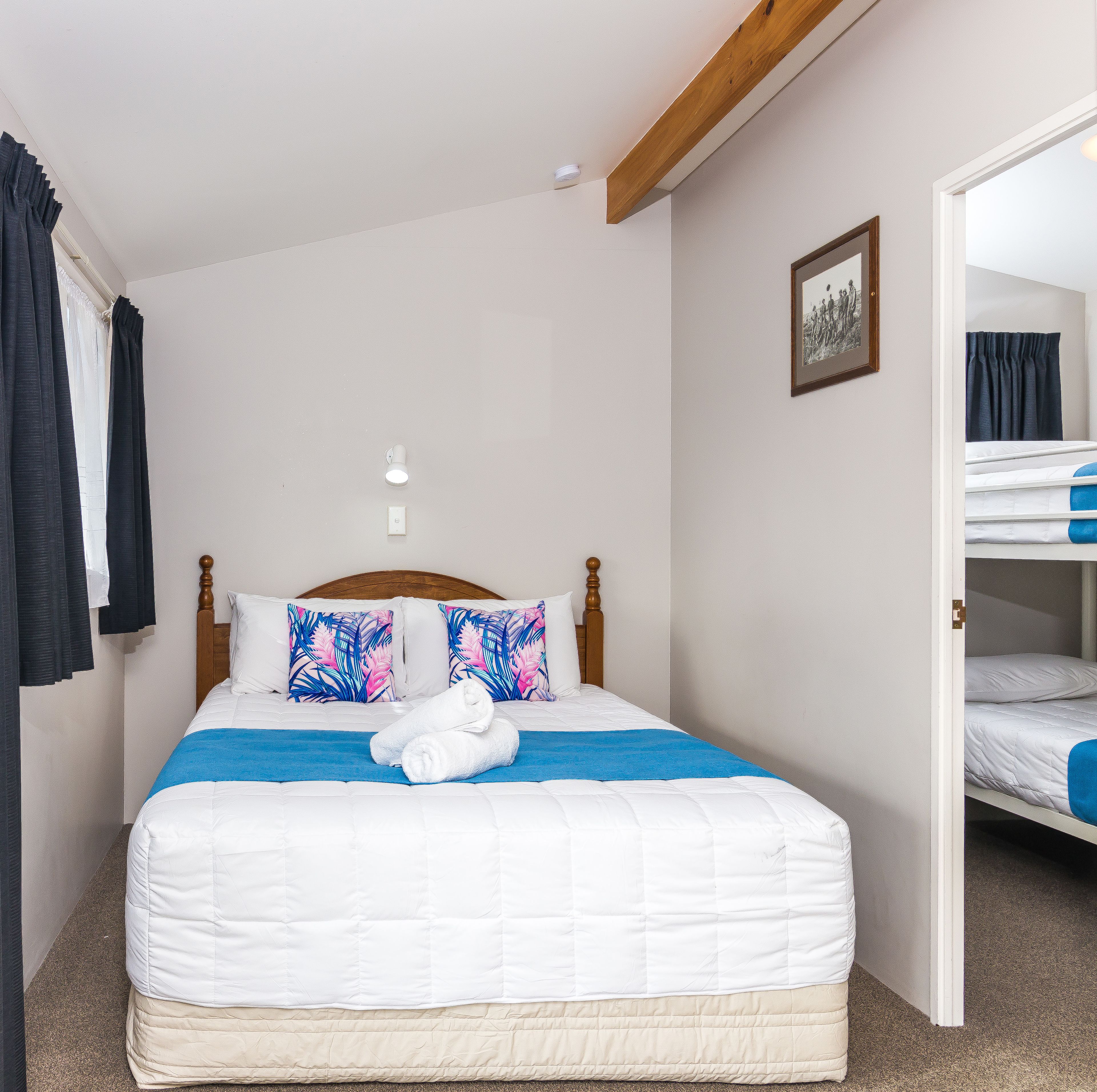 Un lit bien fait, avec une couverture bleue et deux oreillers, une tête de lit en bois et un petit luminaire blanc au plafond.