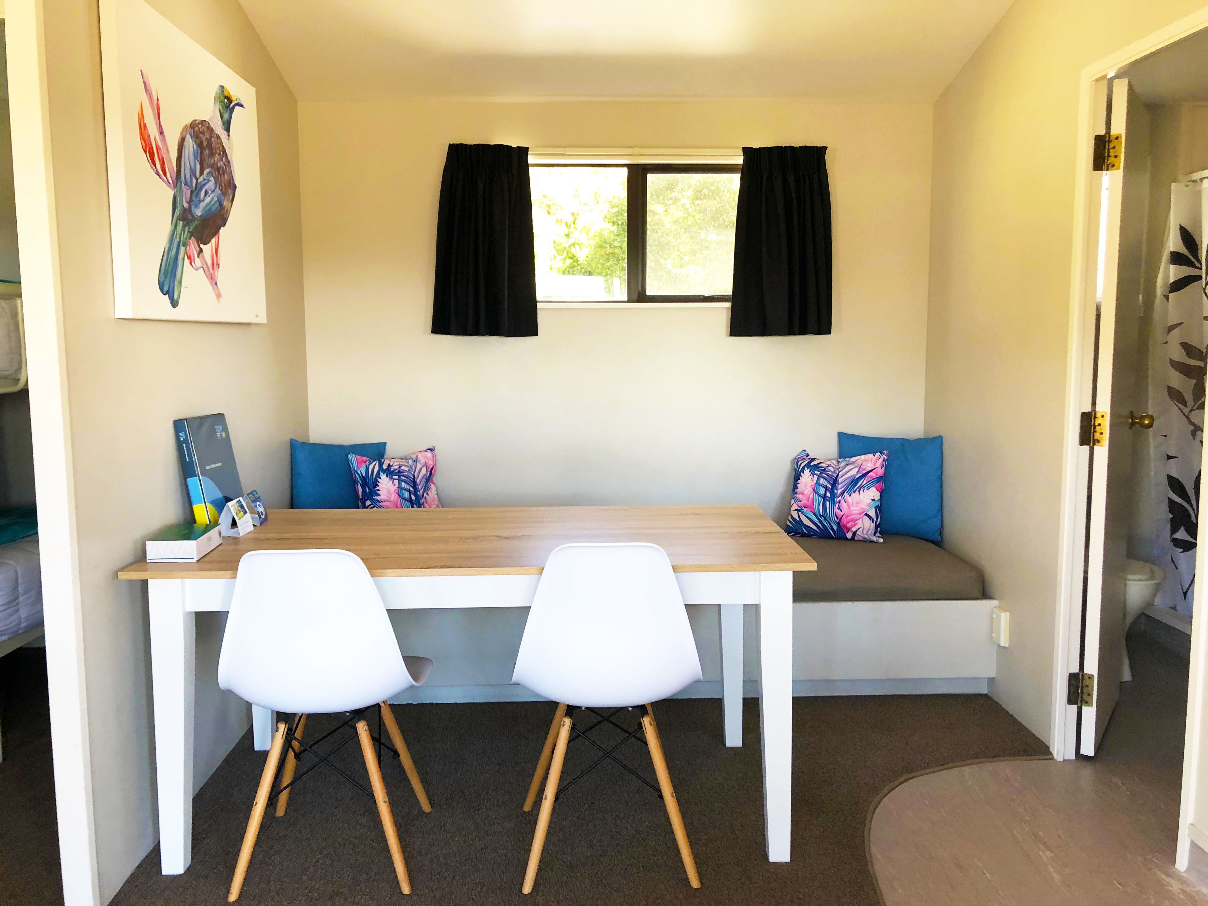 Quarto compacto com mesa de madeira e duas cadeiras brancas, um banco com almofadas azuis, janela com cortinas pretas e porta que dá para um banheiro.