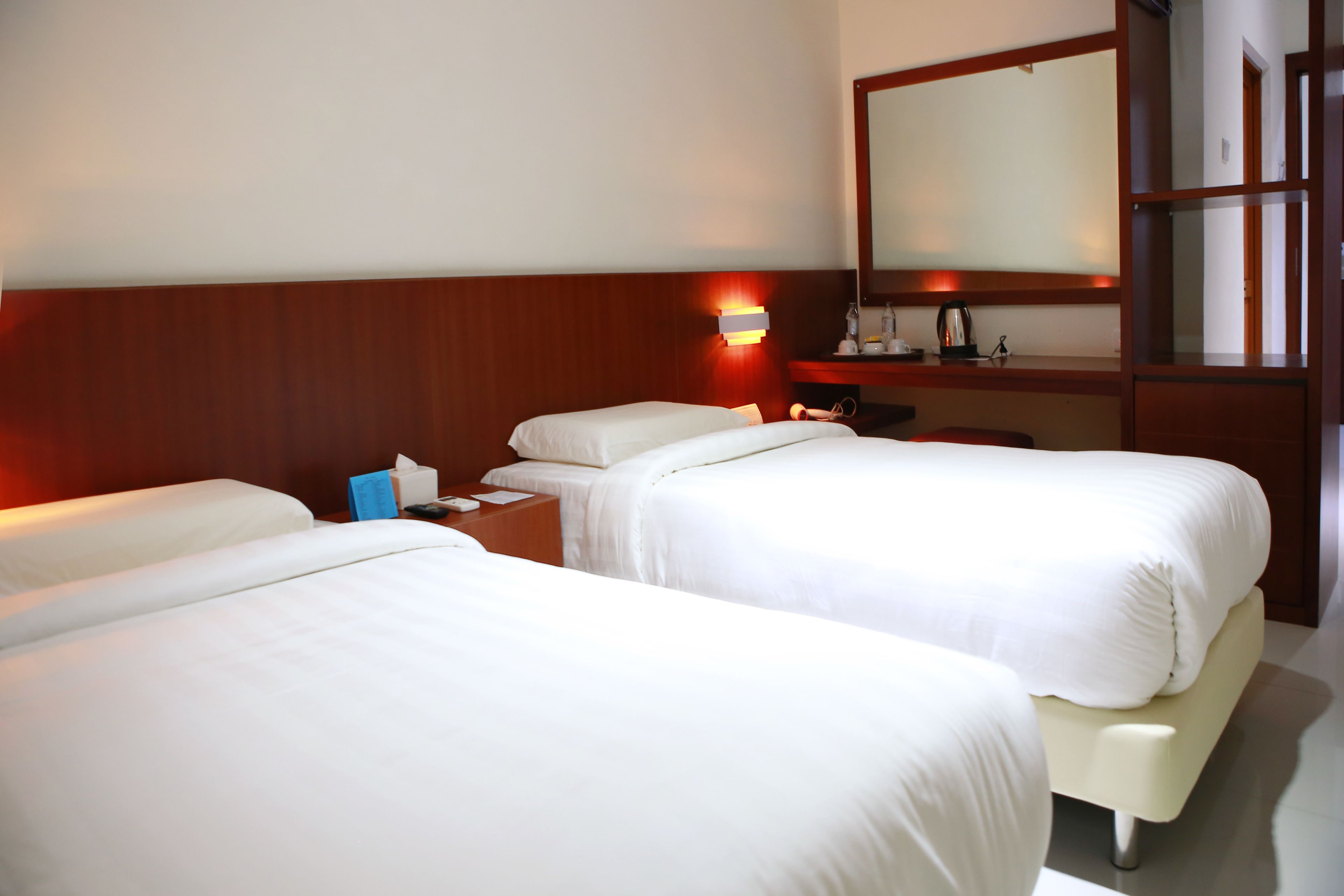 Habitación de hotel con dos camas individuales, cabecera de madera, mesita de noche con lámpara, espejo y estante pequeño con artículos de aseo.