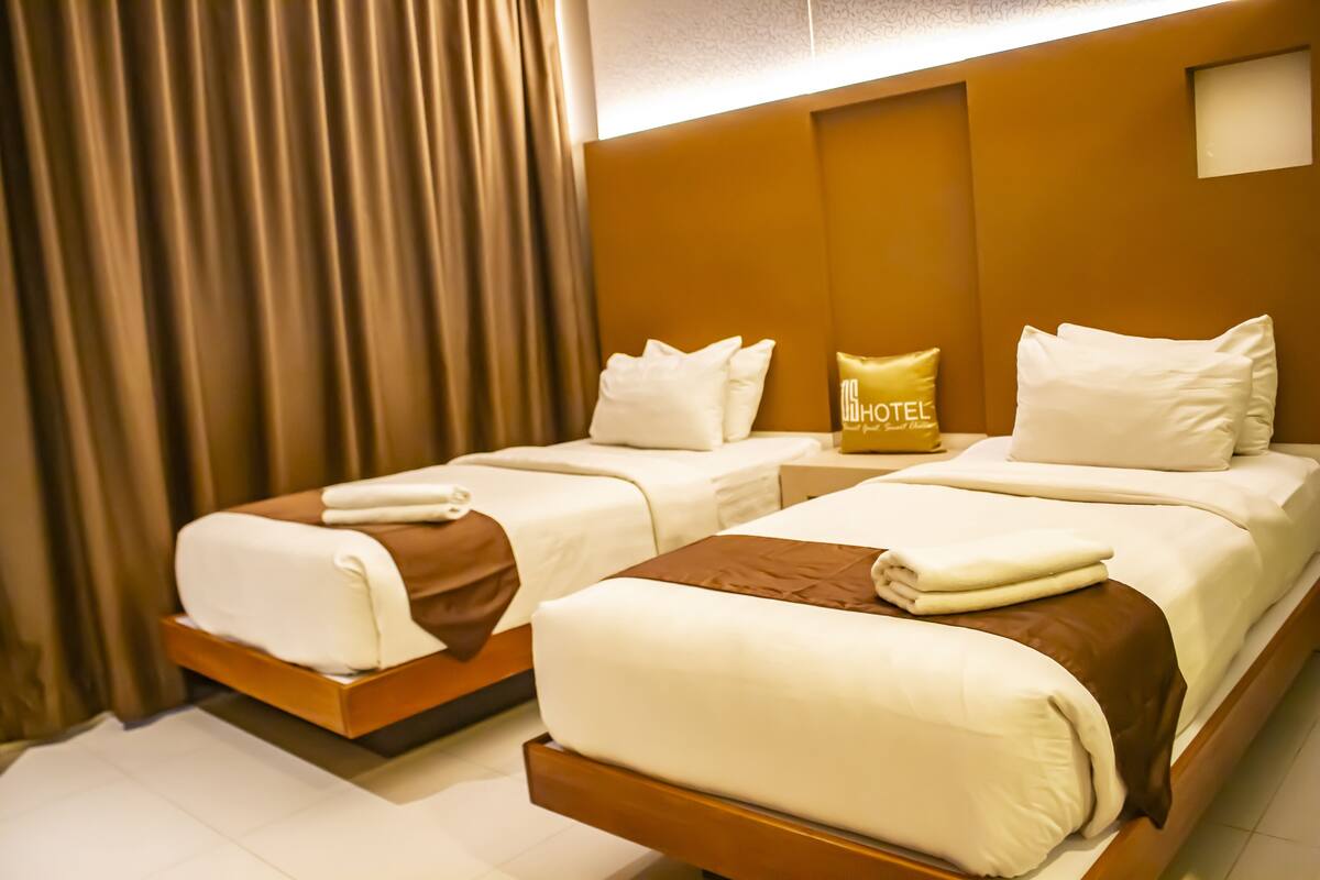 A hotel room with two beds, each with white linens and brown headboards. A 'HOTEL' sign is visible on the wall.