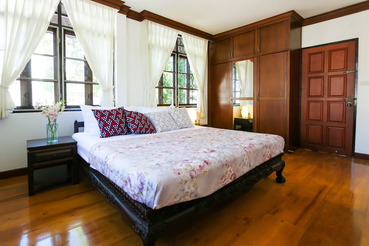Un dormitorio con una cama grande, suelo de madera, una mesita de noche, un aparador y una ventana con cortinas.