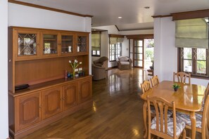 A spacious dining area with a wooden table and chairs, a wooden cabinet with glass doors, and a view of a garden through large windows. - Villa de Nimman (Chiang Mai)