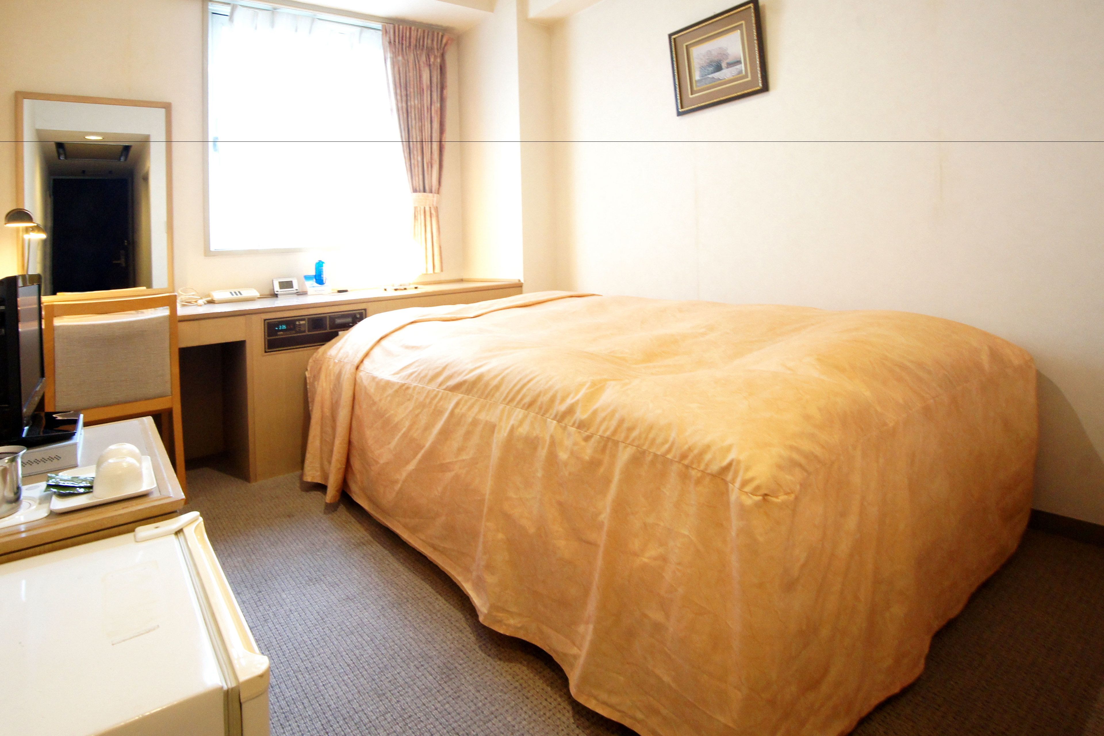 A hotel room with a bed, a desk, a chair, a mirror, a window with curtains, and a framed picture on the wall.