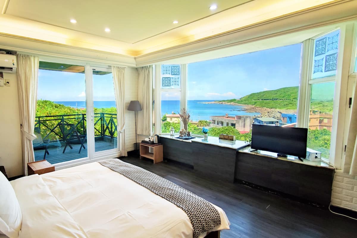 A hotel room with a large bed, a TV, and a balcony with a view of the sea.