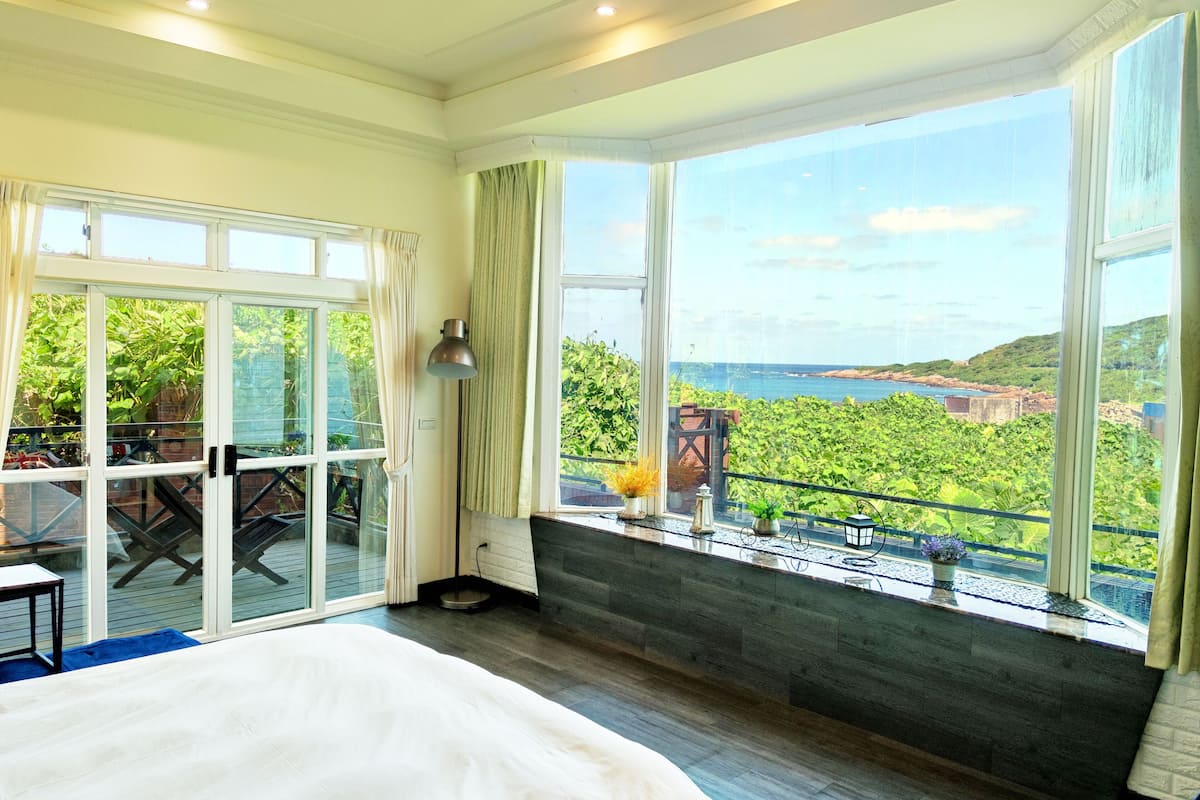 A bedroom with a large bed, a balcony with a view of the sea, and a sliding glass door leading to an outdoor area.