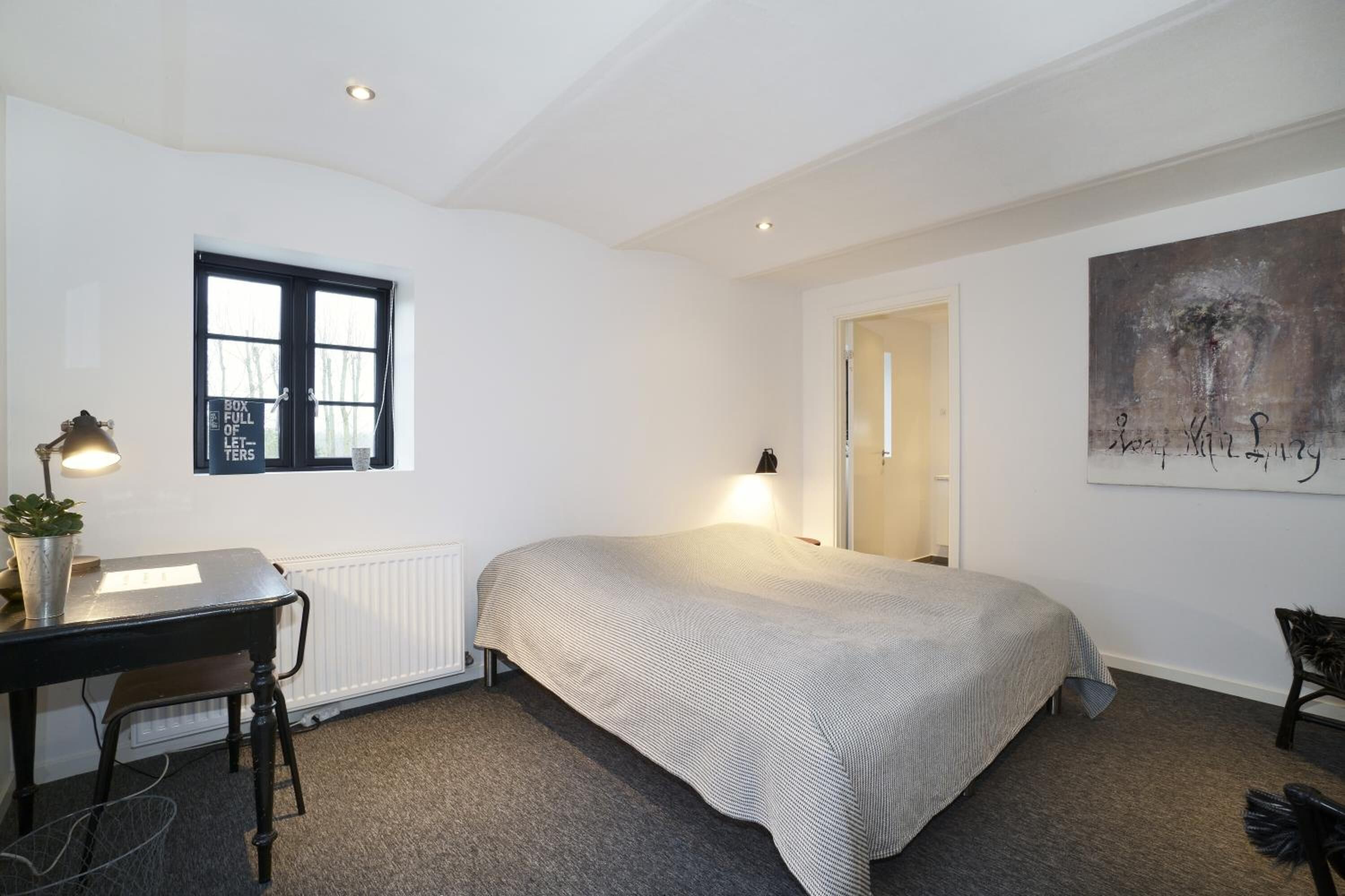 Une chambre à coucher avec un lit, un bureau doté d’une lampe, un radiateur, une fenêtre et un tableau accroché au mur.