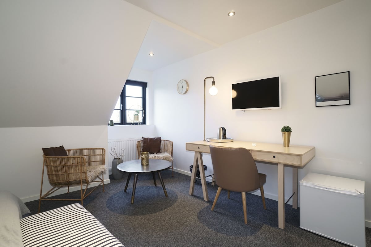 A compact room with a bed, a desk, a chair, a small table, a radiator, a TV, and a window.