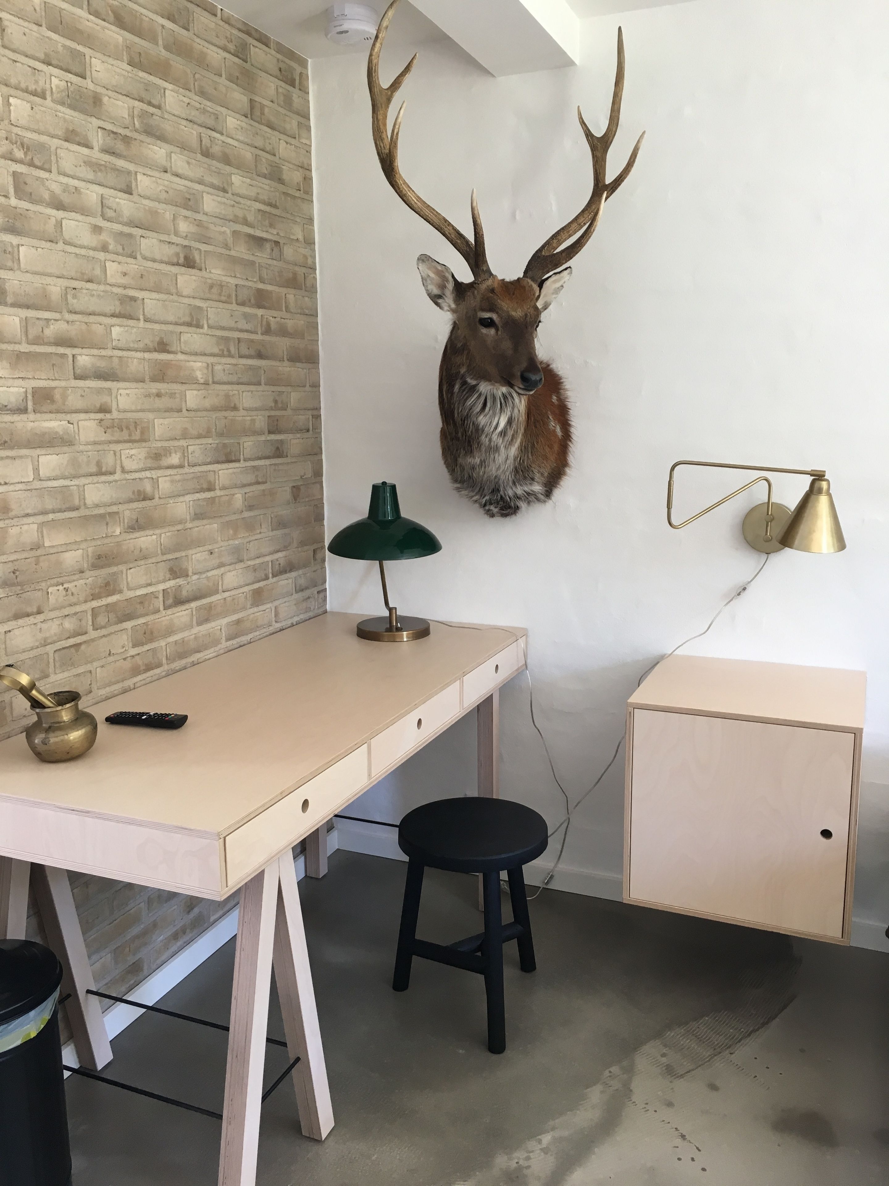 A room with a brick wall, a desk with a lamp, a stool, and a mounted deer head.