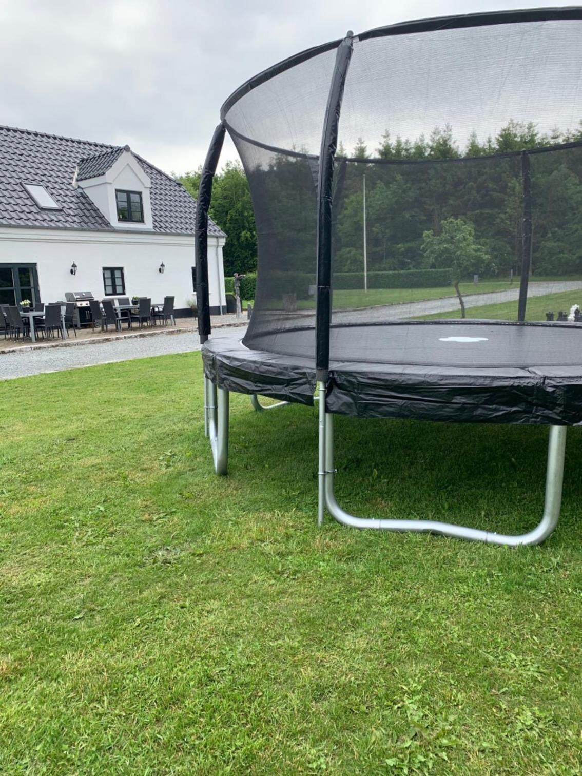 Een trampoline met veiligheidsnet, opgesteld op een gazon voor een huis met pannendak en een terras met tuinmeubilair.