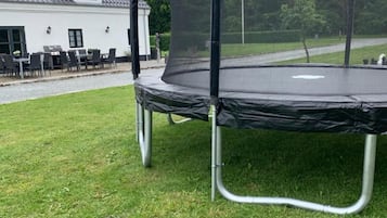 Un trampolino con rete di sicurezza, posizionato su un prato verde davanti a una casa con tetto in tegole e patio con mobili da esterno.