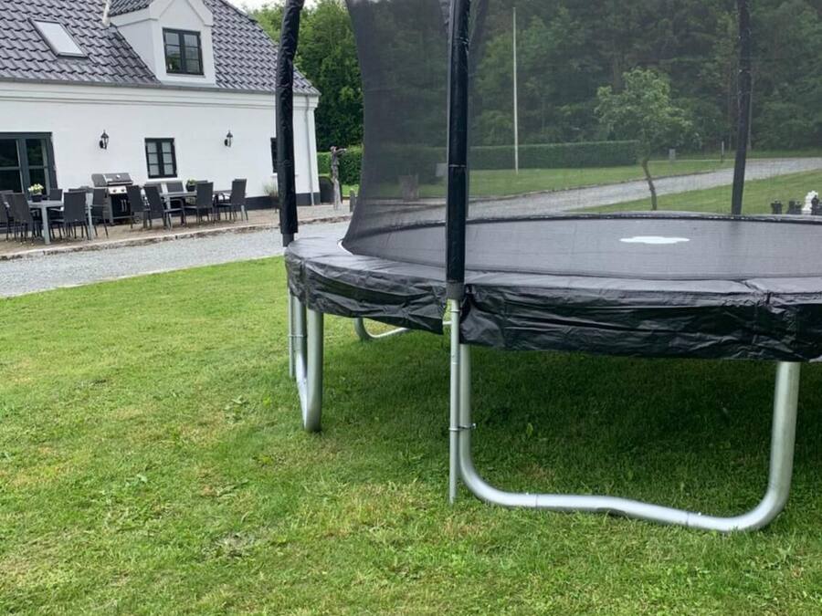 Un trampolino con rete di sicurezza, posizionato su un prato verde davanti a una casa con tetto in tegole e patio con mobili da esterno.