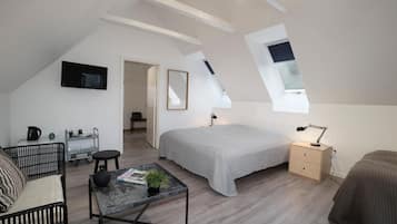 Ein modernes, helles Zimmer mit schrägen Wänden, einem Bett mit grauer Bettdecke, einem kleinen Tisch mit einer Pflanze, einem schwarzen Sofa und einem Nachttisch mit einer Lampe.