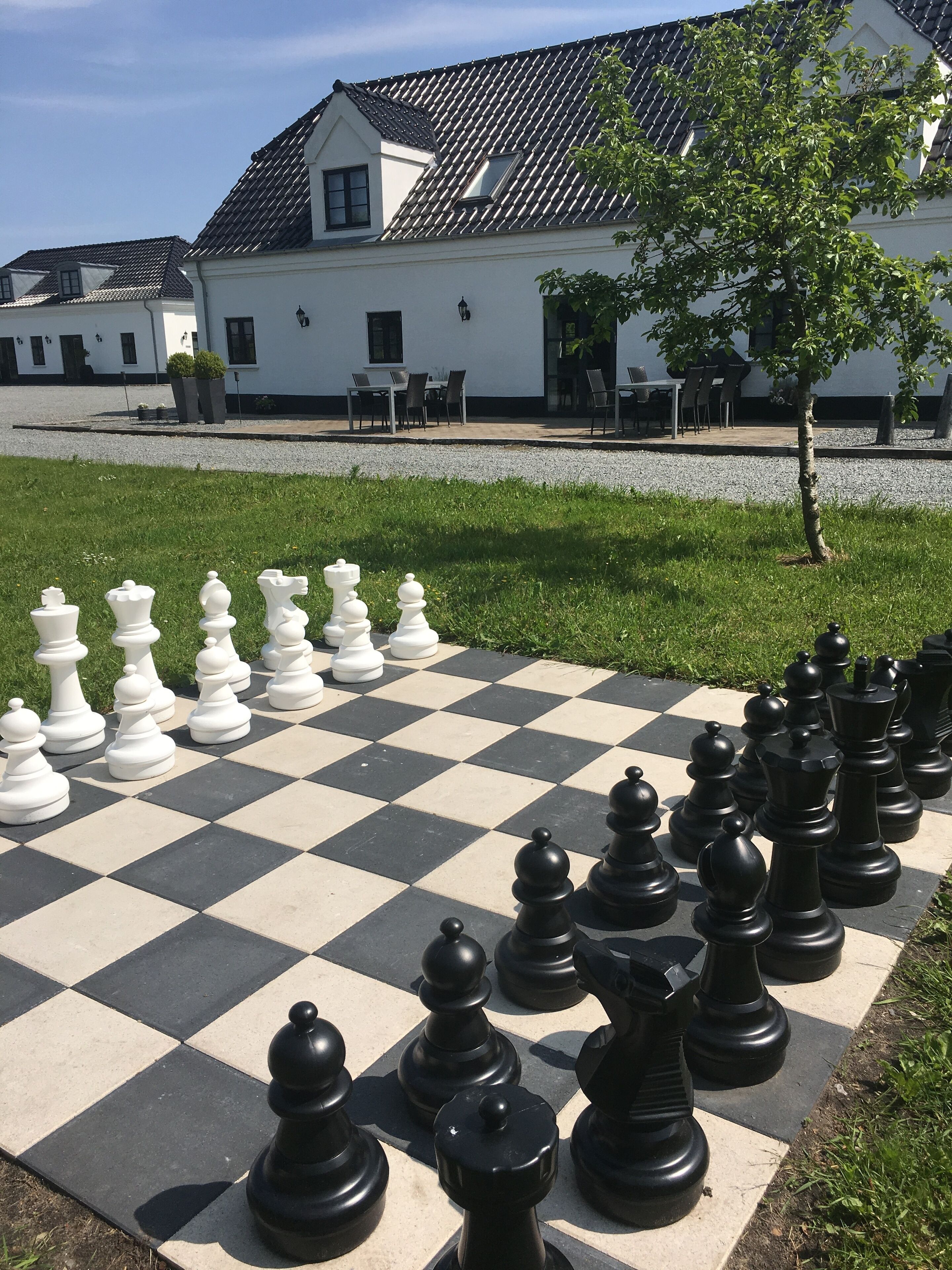 A large outdoor chessboard set up in front of a building with a tiled roof and white walls.