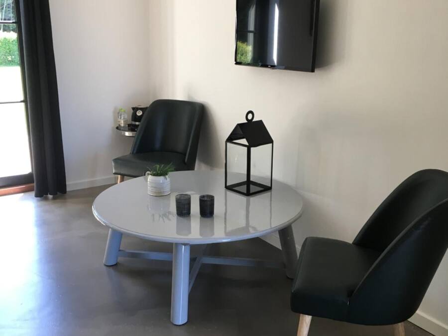 A modern living room with a round glass table, two black chairs, a black floor lamp, and a flat-screen TV mounted on the wall.