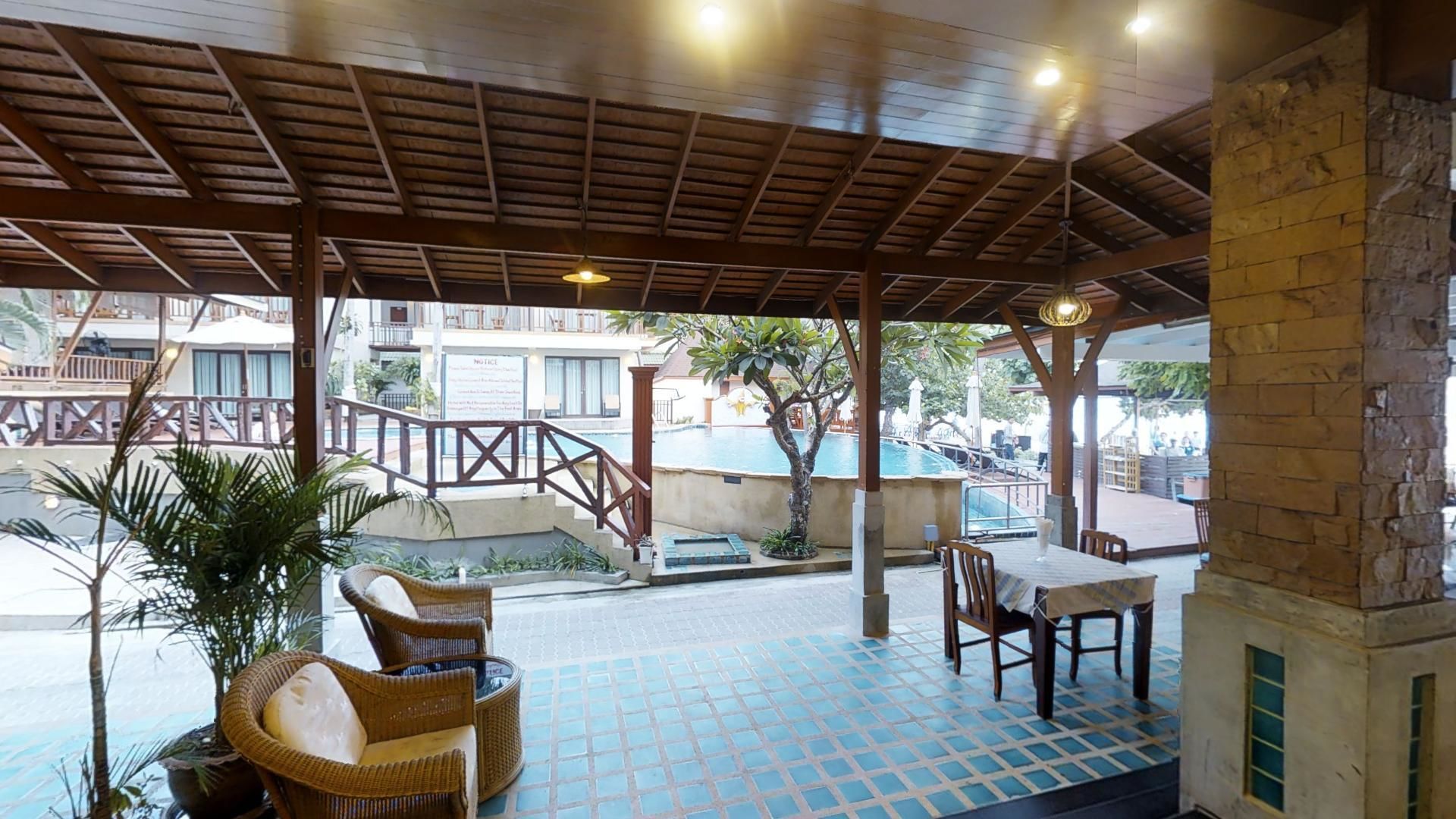 Un espacio de estar al aire libre techado con mobiliario de mimbre, piscina y una escalera que conduce a un edificio.
