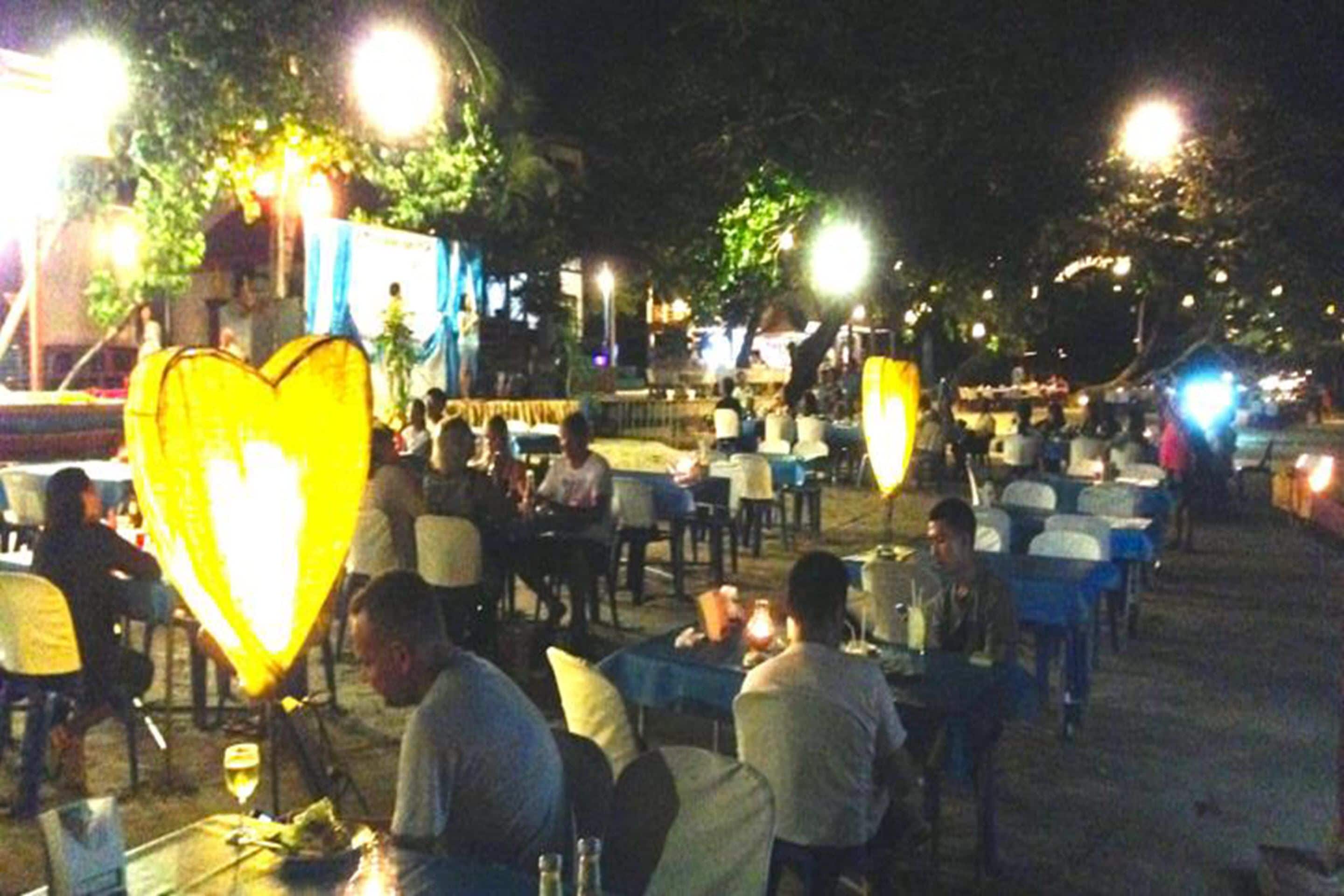 An outdoor dining area with tables and chairs, surrounded by trees and street lamps.
