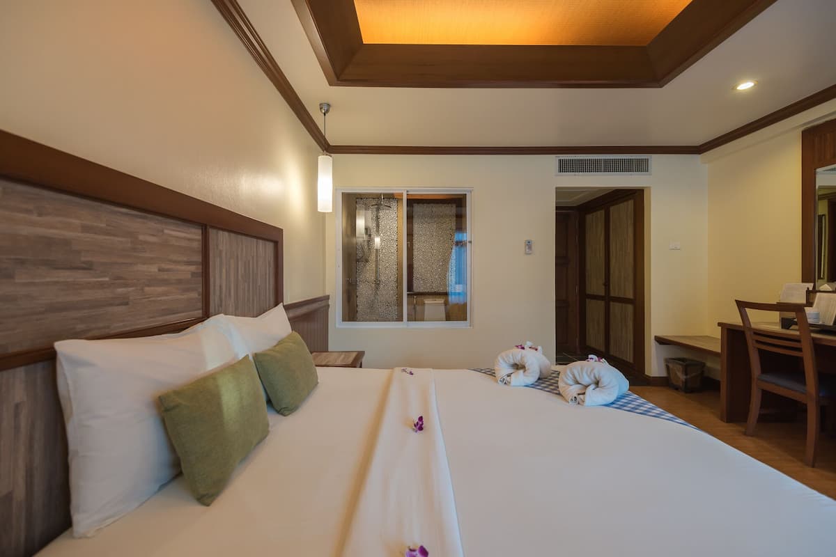 A hotel room with a large bed, a wooden headboard, a window with patterned curtains, a desk, and a chair.
