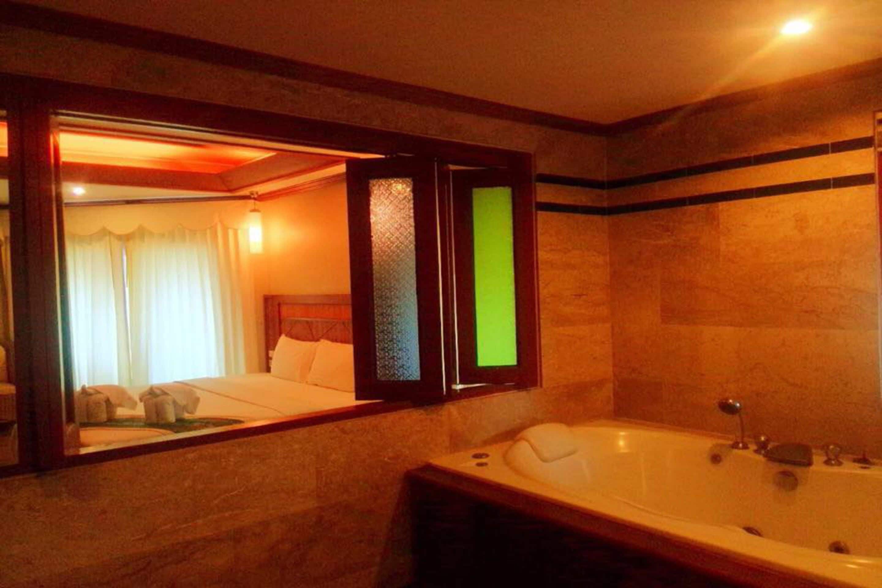 Ein Hotelbad mit einer großen Badewanne, einem Spiegel, der einen Raum mit einem Bett und Vorhängen widerspiegelt, und einer gefliesten Wand.