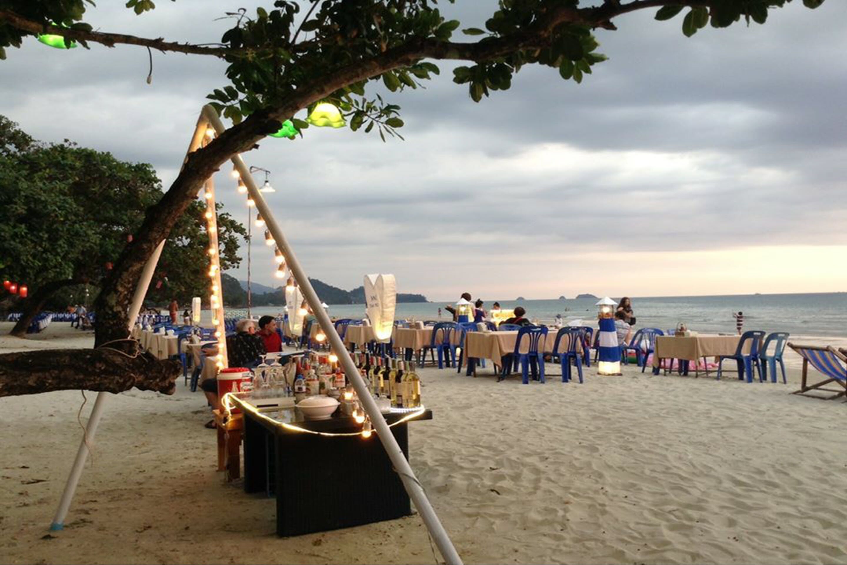 An outdoor dining area with tables and chairs arranged on a sandy beach, overlooking the sea.