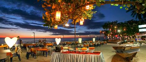 A beachside restaurant with tables and chairs, a buffet setup, and a boat on the sand.