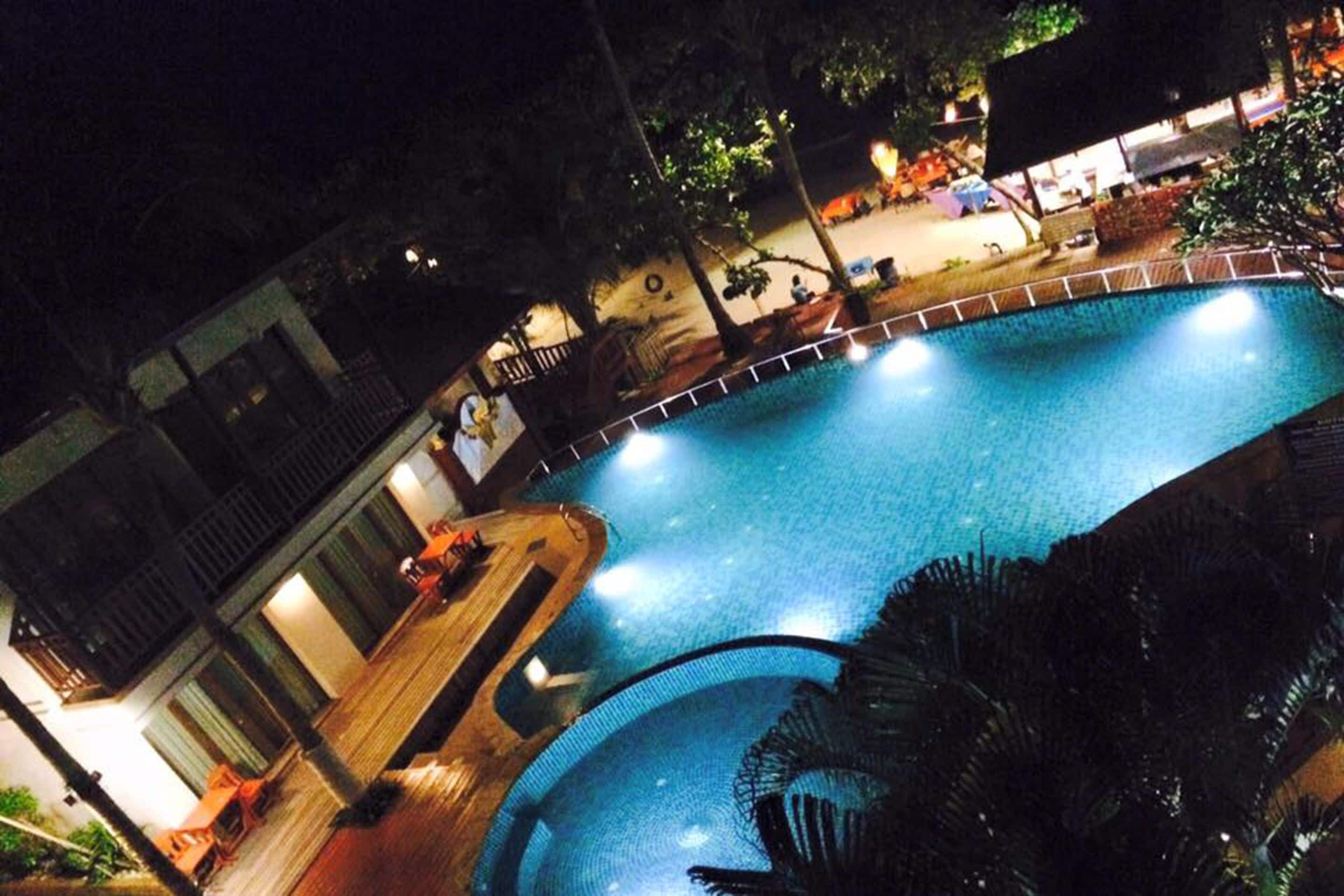 A nighttime view of a swimming pool with a wooden deck and palm trees.