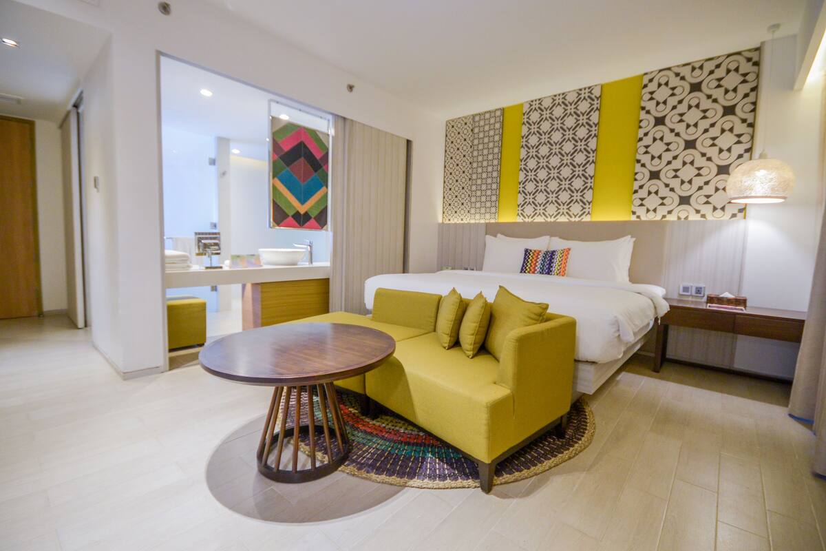 A hotel room with a bed, a yellow sofa, a round table, and a patterned wall.