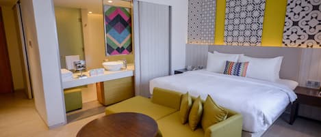 A hotel room with a bed, a sofa, a small table, a bathroom with a sink and mirror, and a patterned wall.