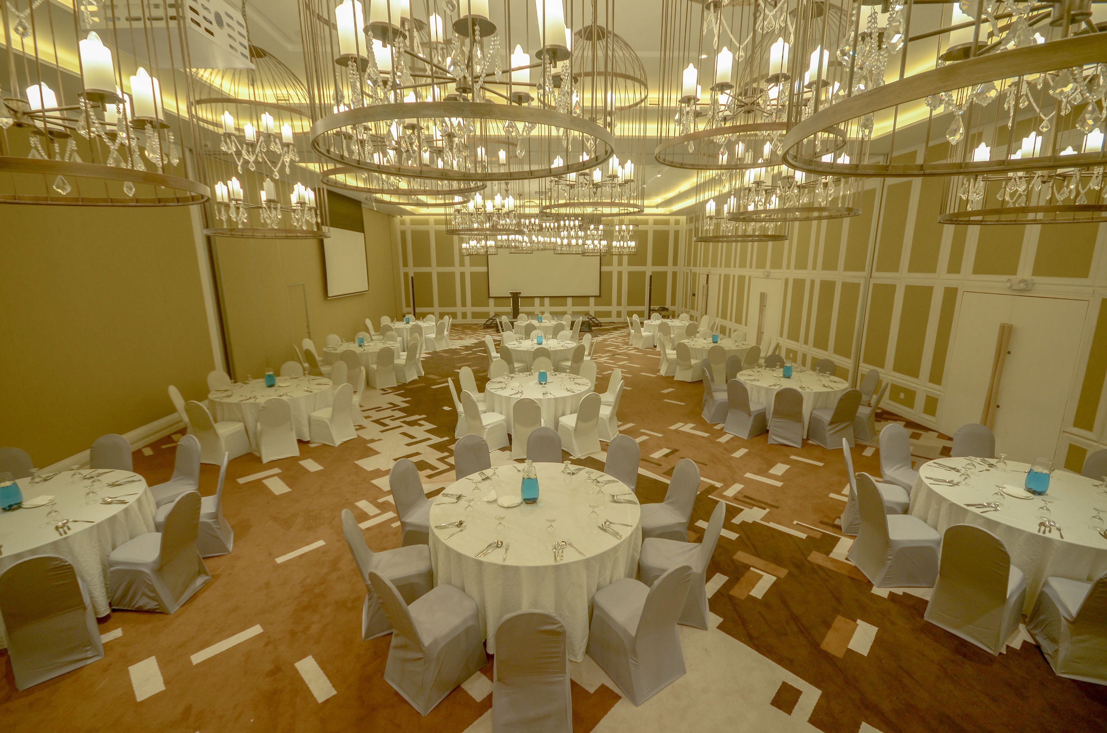 Un salón de banquetes con varias mesas redondas, sillas blancas y una pantalla grande.