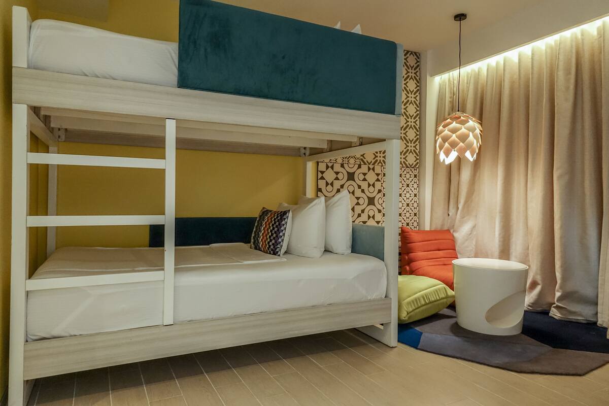 A bunk bed with a ladder, a patterned wall, a hanging light fixture, and a window with curtains.