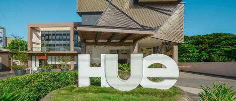 A modern building with a large 'HUE' sign in the foreground, surrounded by greenery and a clear sky.
