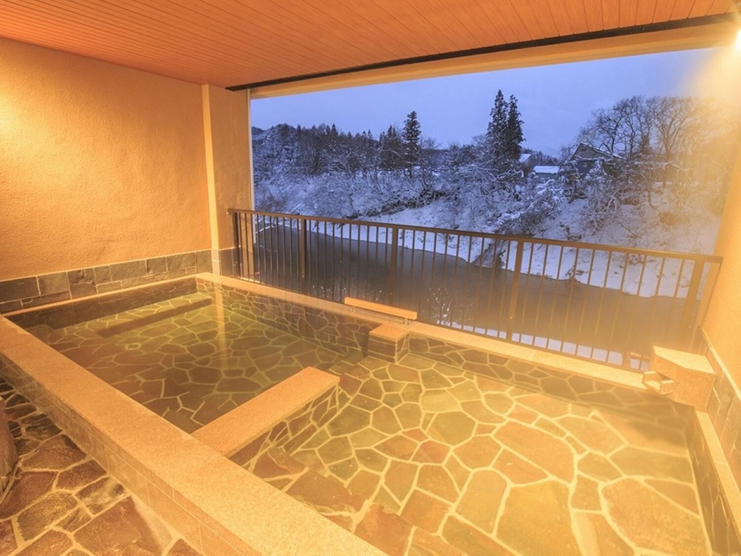 A stone-floored hot tub with a wooden ceiling and a view of a snowy landscape through a balcony railing.