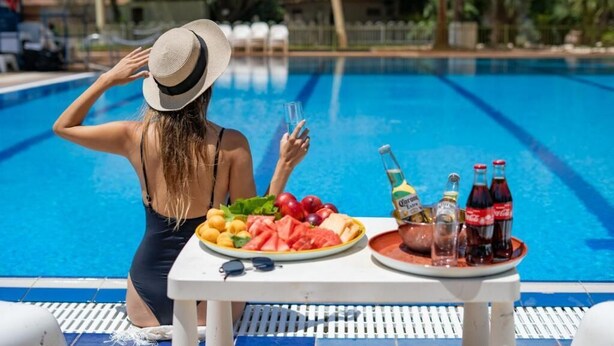 Una scena a bordo piscina con un tavolo su cui ci sono frutta, bottiglie e un bicchiere.