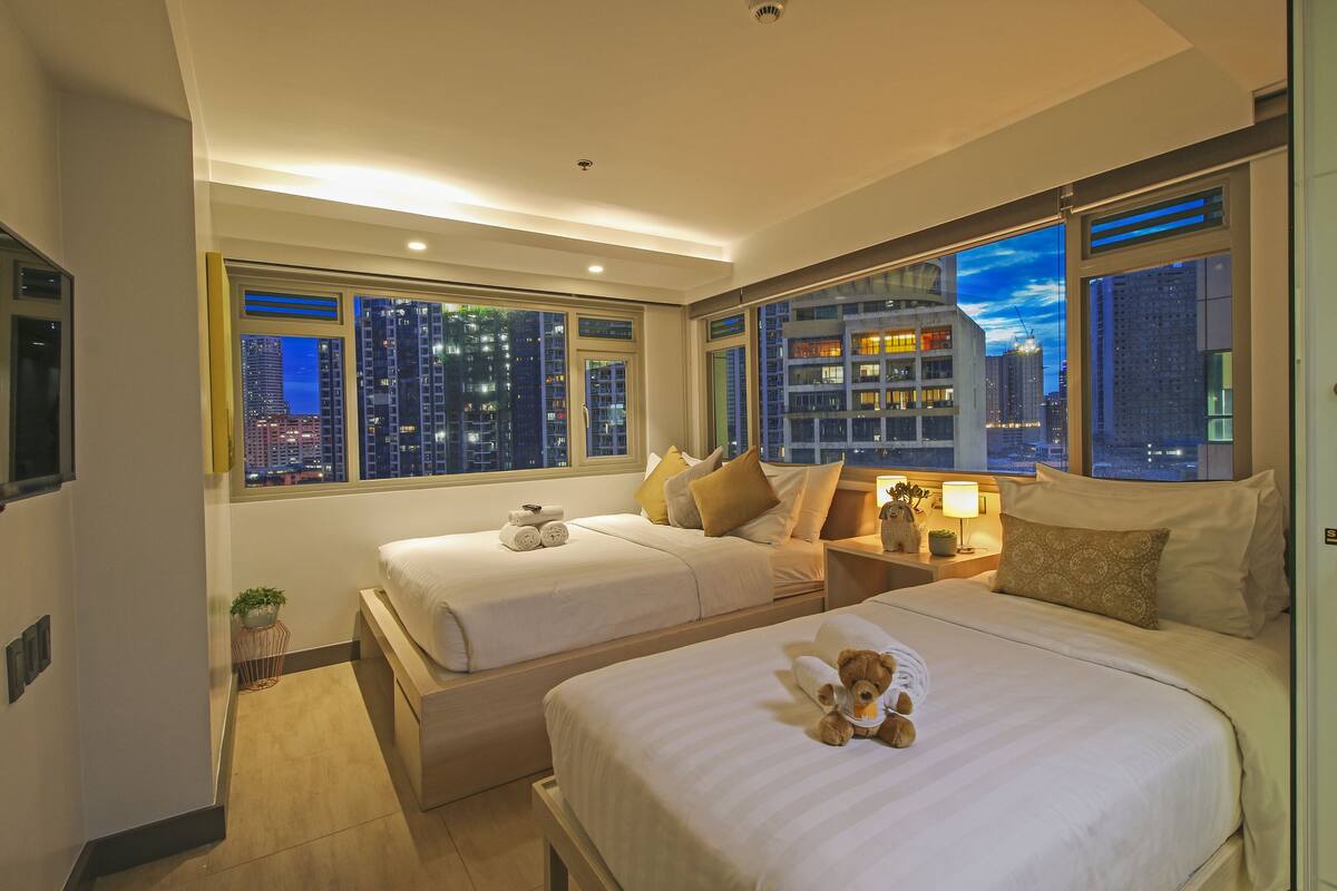 A hotel room with two beds, a nightstand, a lamp, and a view of the cityscape at night.