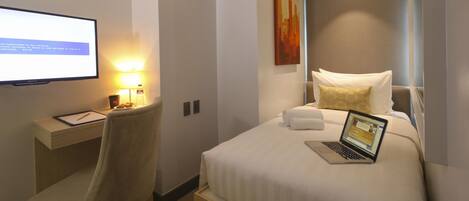 A hotel room with a bed, a desk with a laptop, a chair, a lamp, and a TV on the wall.