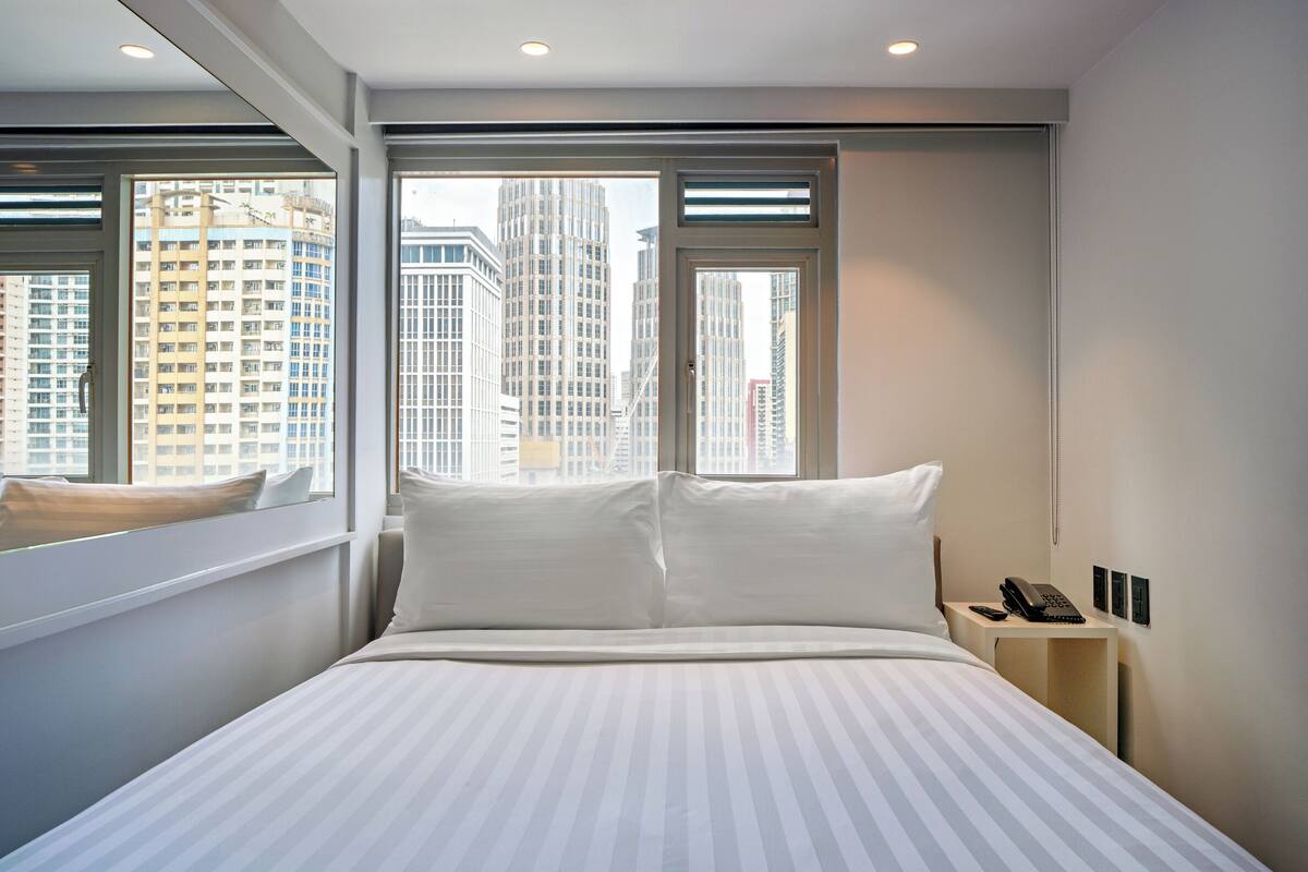 A modern hotel room with a large bed, a bedside table with a phone, and a window offering a city view.