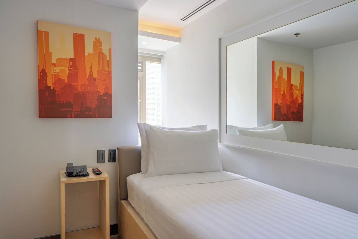 A hotel room with a bed, a bedside table with a phone, a mirror, and a cityscape painting on the wall.