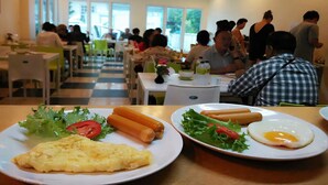 Un restaurante con varias mesas y sillas, personas comiendo y platos de comida que incluyen huevos, perritos calientes y ensaladas.