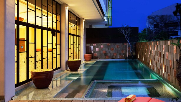 A modern outdoor pool area with a glass enclosure, wooden deck, and red lounge chairs. - Grand Livio Kuta Hotel (Legian)