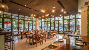 A spacious restaurant with wooden tables and chairs, large windows, and hanging pendant lights. - Grand Livio Kuta Hotel (Legian)